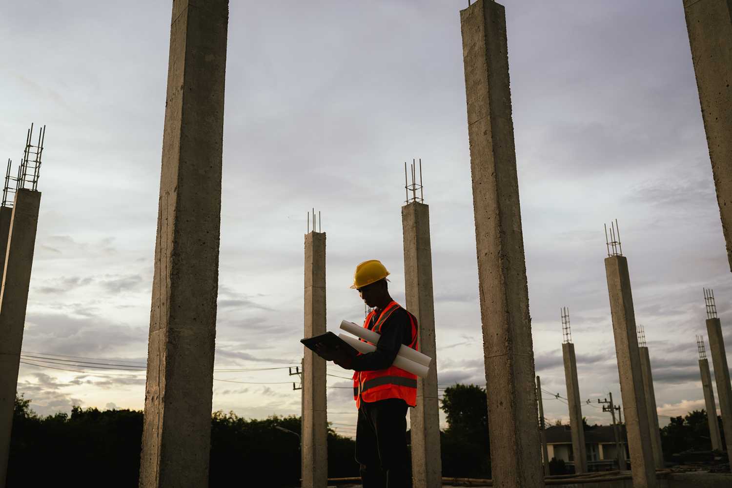 Silhouette of a construction worker holding blueprints and using a phone at a building site during sunset, symbolizing planning, progress, and evening work on-site.