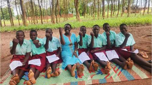 A researcher sits outdoors on a mat with seven girls in school uniforms in northern Uganda. All raise their fists toward the camera.