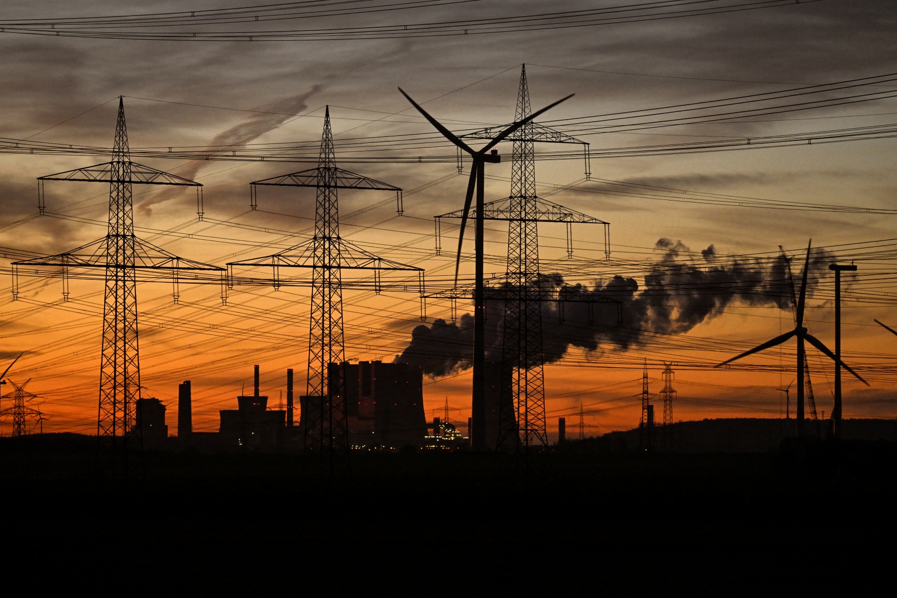 This picture taken at sunset on November 12, 2025 shows wind turbines and power poles as fumes rise from the cooling towers of the lignite-fired power station operated by German energy giant RWE near Neurath, western Germany.
