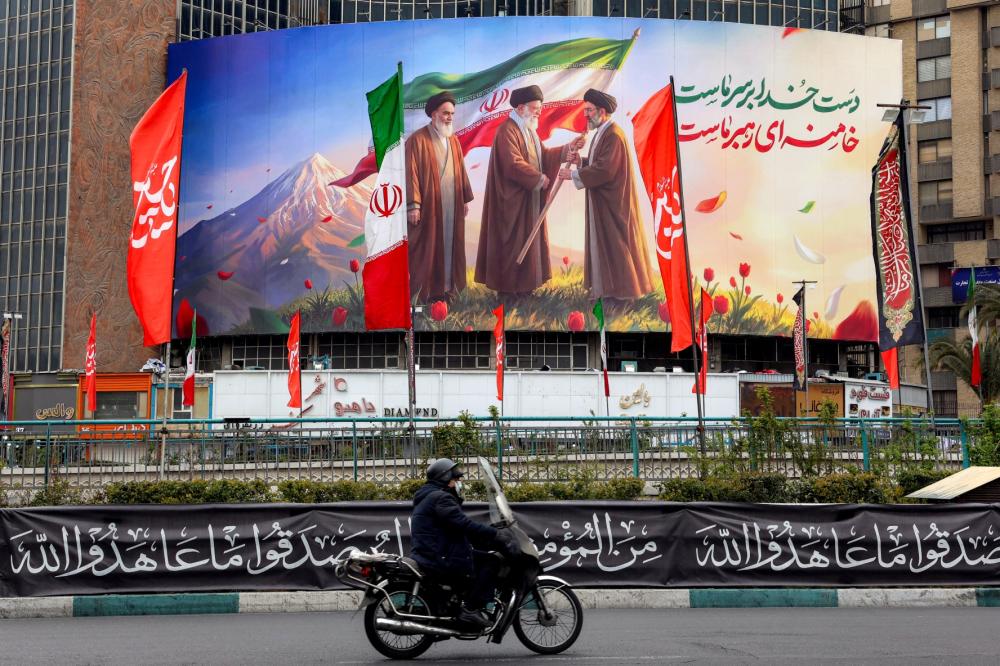 A man rides a motorcycle past a banner displayed at Valiasr Square in central Tehran on March 10, 2026, depicting Iran's late supreme leader Ayatollah Ruhollah Khomeini (L) watching as his successor the late Ayatollah Ali Khamenei (C) hands over a national flag to his son and new supreme leader Mojtaba Khamenei.