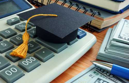Graduation cap on top of a calculator next to stacks of money, a pen, and notebooks.