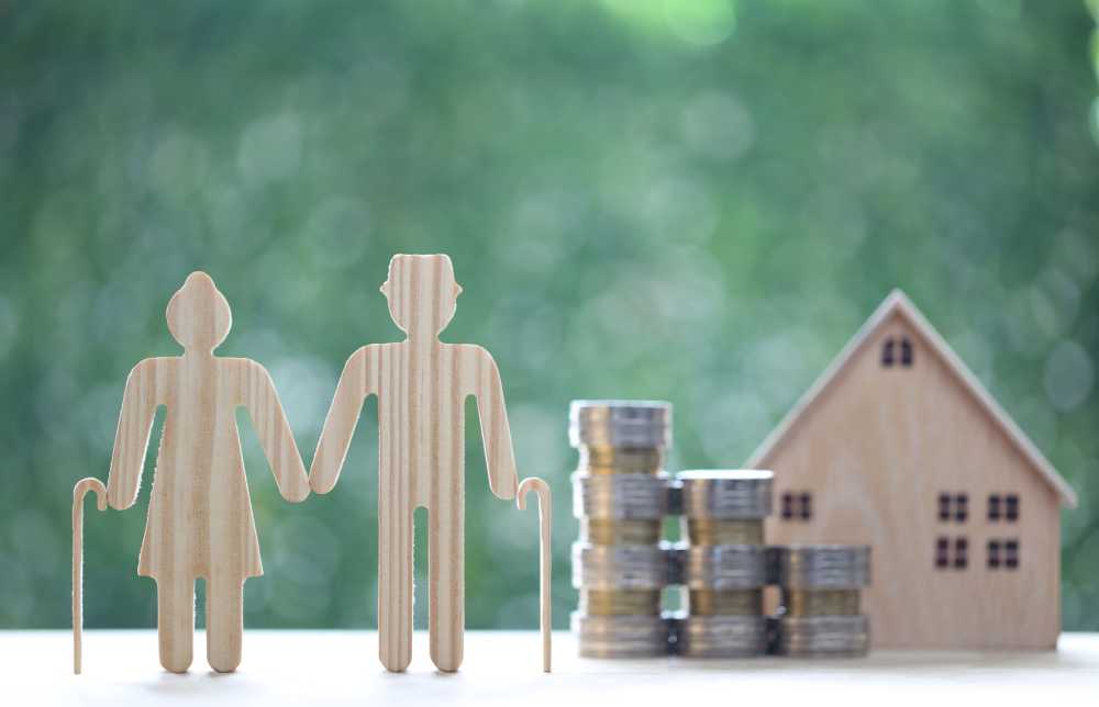 Wooden cutouts of a senior couple and a house next to a stack of coins, against a green background.