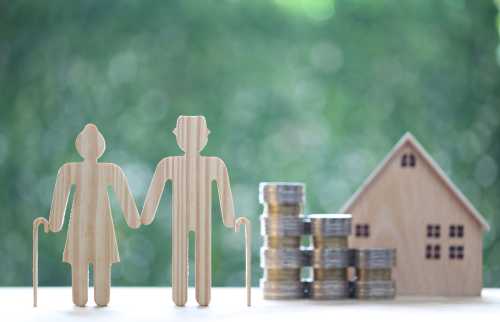 Wooden cutouts of a senior couple and a house next to a stack of coins, against a green background.