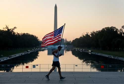 WASHINGTON, DC - JULY 03: Pete Folch carries an American flag during a morning run past the Reflecting Pool as the city prepares for July 4th festivities on July 3, 2026, in Washington, D.C.
