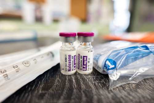 Two vials of intramuscular naloxone 0.4mg/mL sitting on a wooden table next to a packaged syringe and some other supplies.