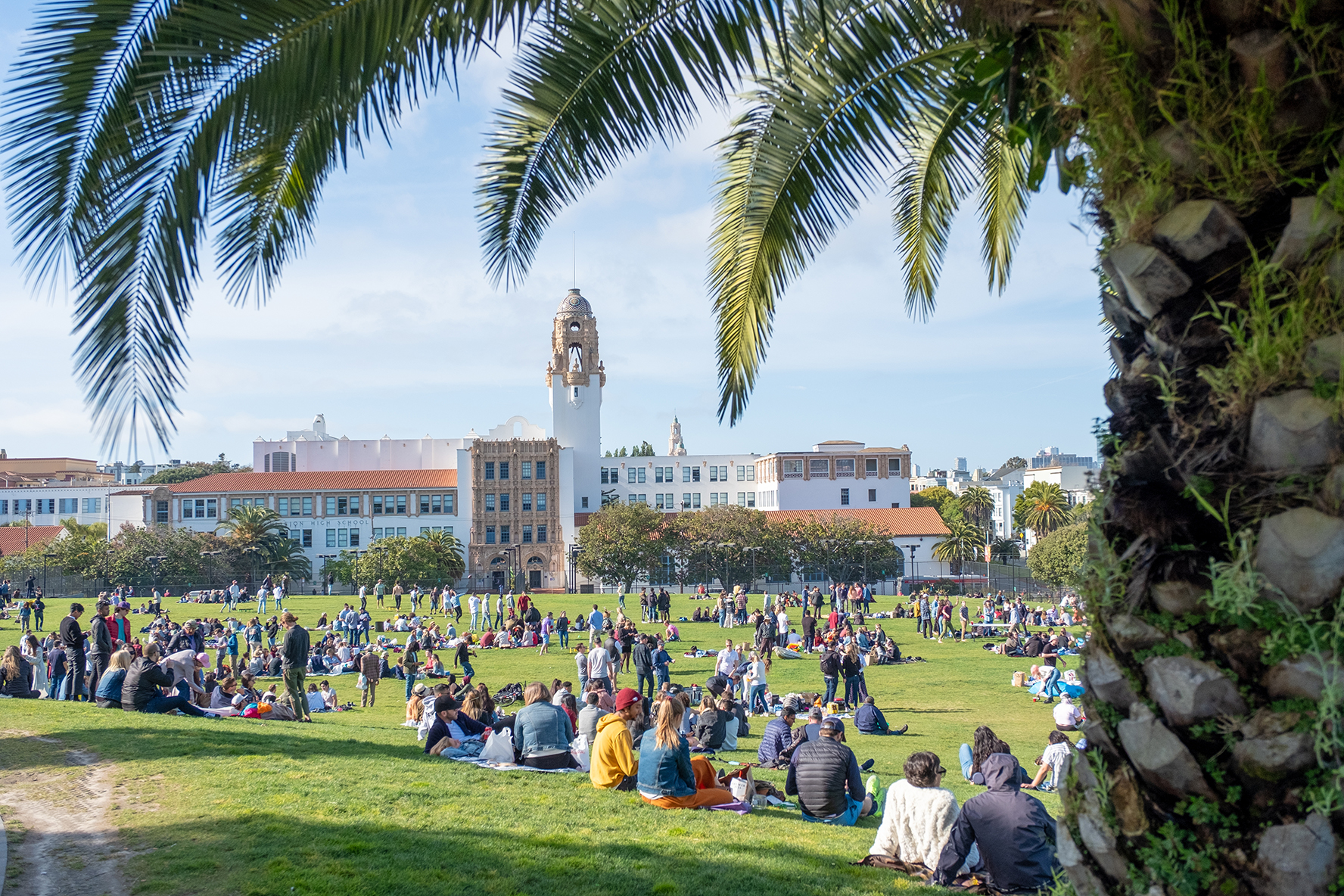People sitting beneath tall palm trees enjoying a sunny picnic at Mission Dolores Park with the tower of Mission High School visible, San Francisco, CA on May 4, 2019