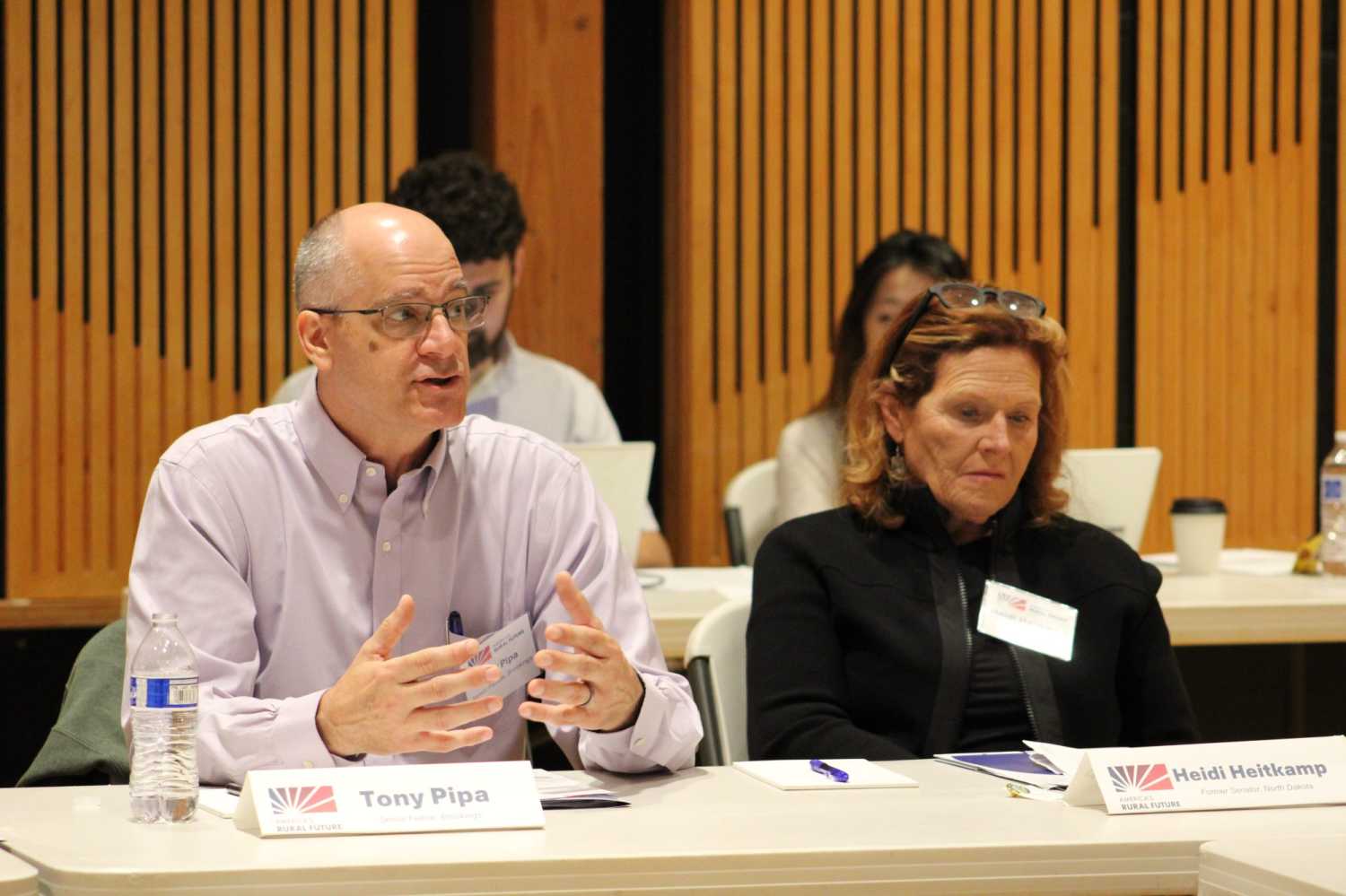 Tony Pipa gestures while asking a question, seated beside Heidi Heitkamp at a table with America's Rural Future nameplates during a Commission site visit.