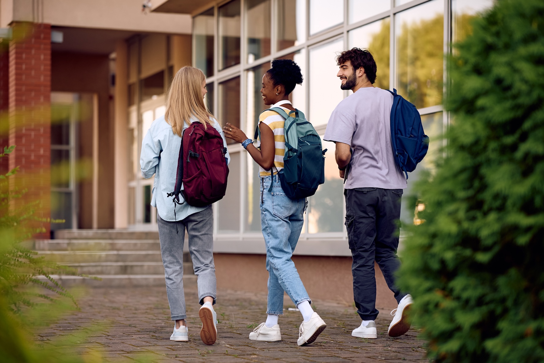 Three students wearing backpacks laugh and walk across a college campus