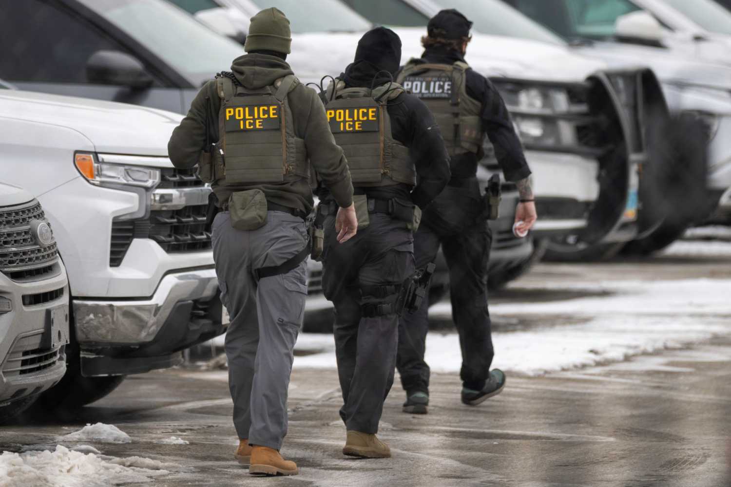 Three men dressed in protective vests marked "Police ICE" walk through a snowy parking lot.