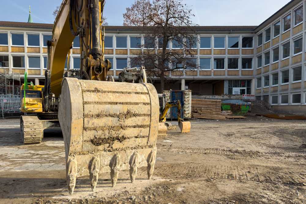 Workers oversee a school construction site as an excavator digs near building materials and other equipment.
